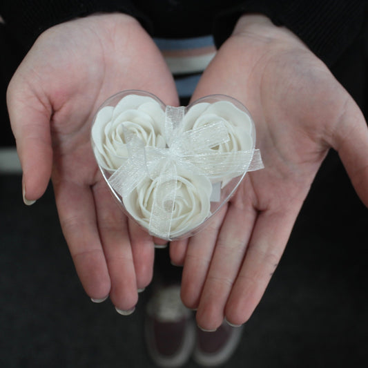 Heart-shaped clear box holding three white rose-shaped soaps tied with a sheer ribbon, cupped in a pair of outstretched palms above a dark floor with visible shoes.