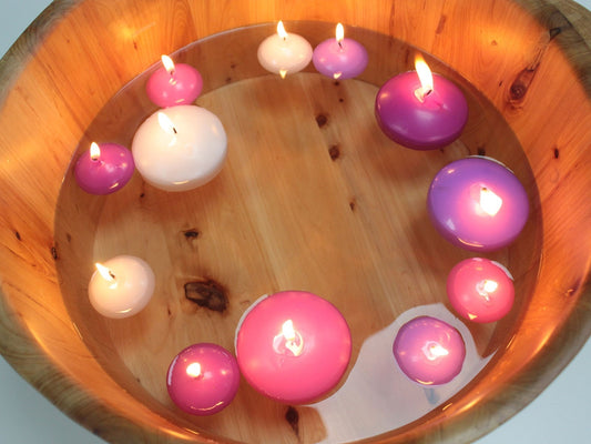 Floating round candles burn while drifting in a wooden bowl of water, various pink, purple and white candles arranged around the bowl's interior, warm reflections on the wood.