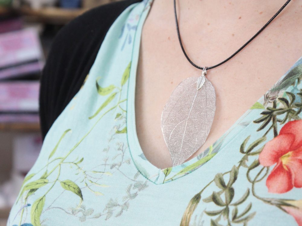 A silver leaf pendant hangs on a black cord around a person's neck, with floral patterns on their light blue V-neck shirt.