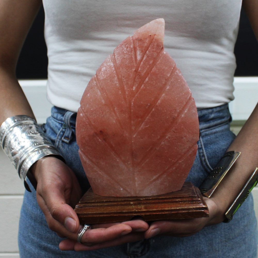 A carved pink leaf-shaped stone is held by a person wearing a white sleeveless top and blue jeans, adorned with silver bracelets, against a blurred background.