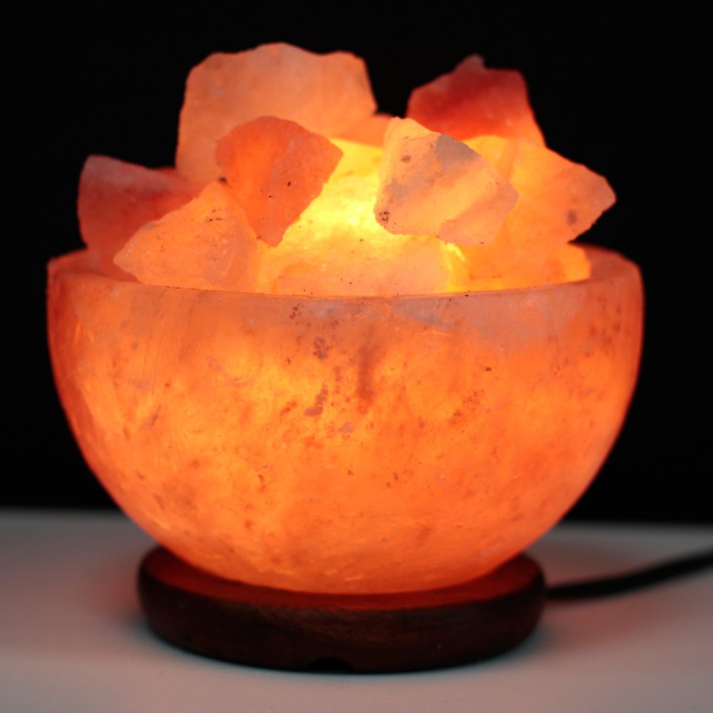 A glowing orange salt lamp bowl, filled with irregularly shaped chunks, illuminates softly in a dark setting, resting on a wooden base.