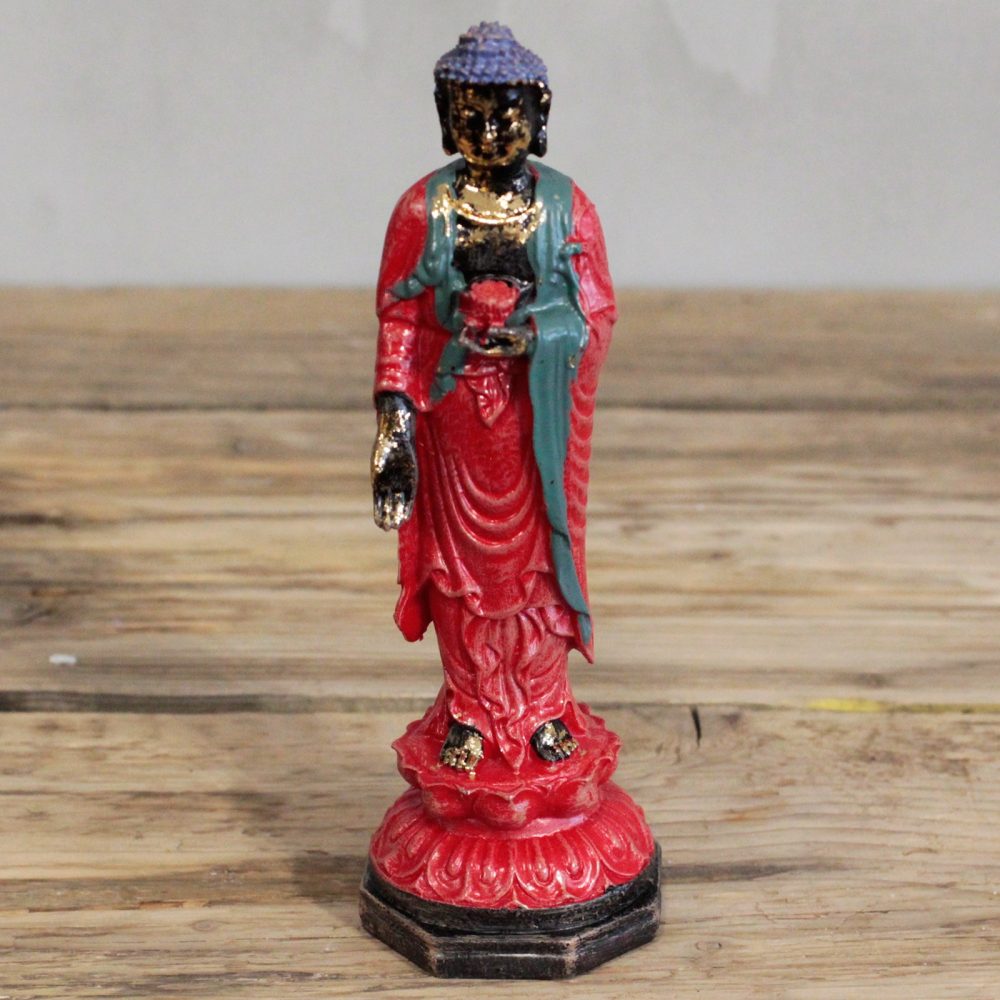 A small, colorful Buddha statue stands on a wooden surface. The statue features red and green robes and is positioned in an upright posture, exuding a serene presence.