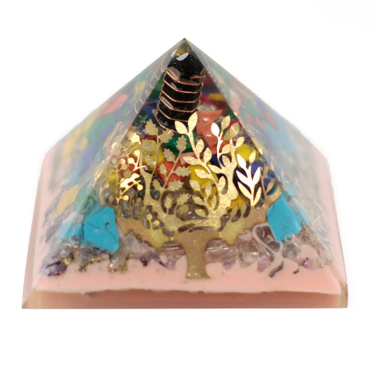 A pyramid-shaped orgonite holds colorful gems, a gold-toned tree design, and a copper coil inside a transparent, pale-pink base. The pyramid is set against a white backdrop.