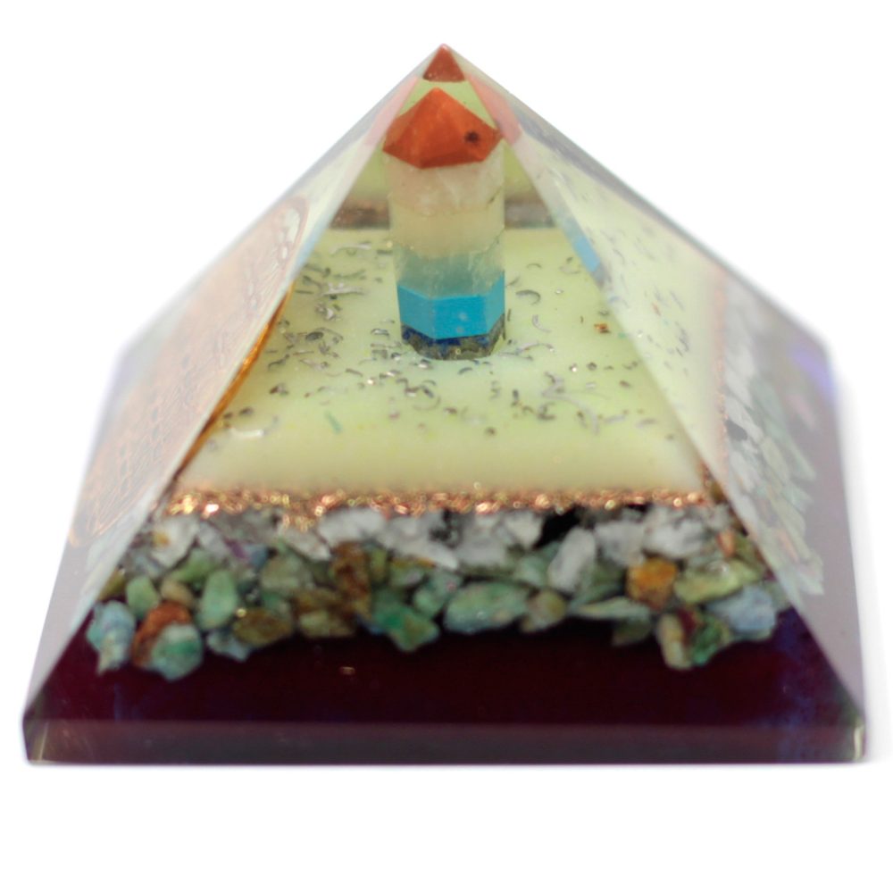 A layered orgonite pyramid sits against a white background; layers include small colorful rocks, followed by metal shavings, a light green layer with glitter, and a stack of colored crystals at the apex.