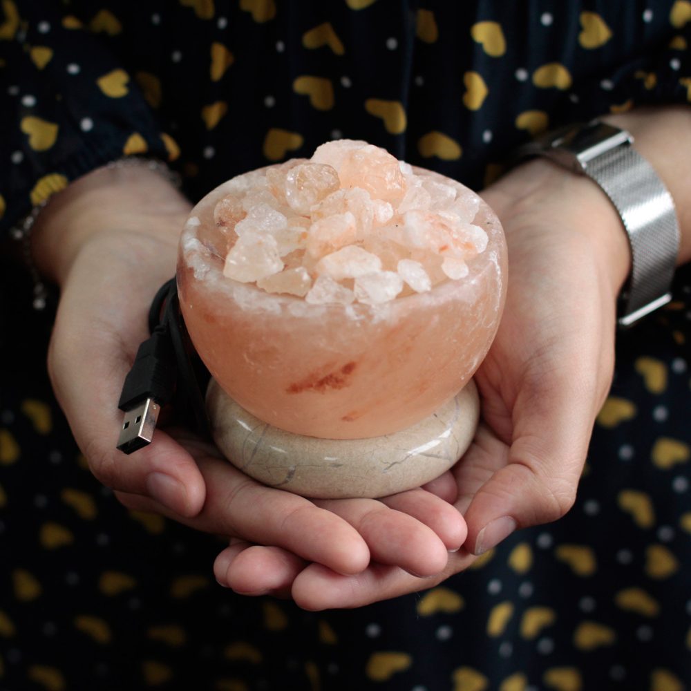 A bowl-shaped Himalayan salt lamp rests in cupped hands, filled with salt crystals, and attached to a USB cable. The person wears a patterned outfit in a dimly lit space.