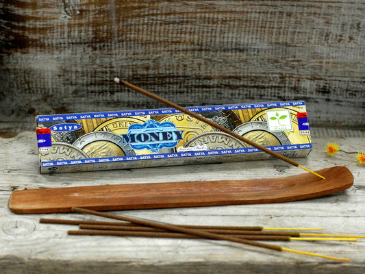 Incense sticks rest on a wooden holder, with a "Satya Money" incense box in the background displaying images of coins. Small yellow flowers are scattered on a rustic wooden surface.