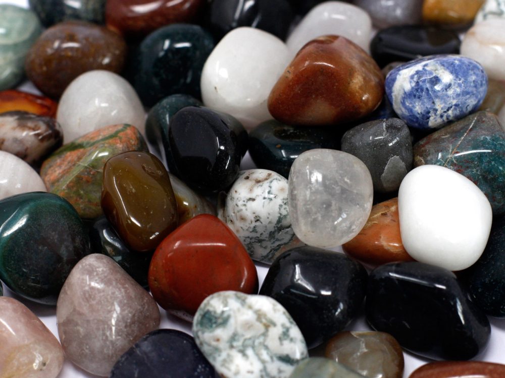 Polished, colorful stones are clustered together, with many shapes and sizes. The shiny rocks include shades of white, red, black, brown, gray, and blue.