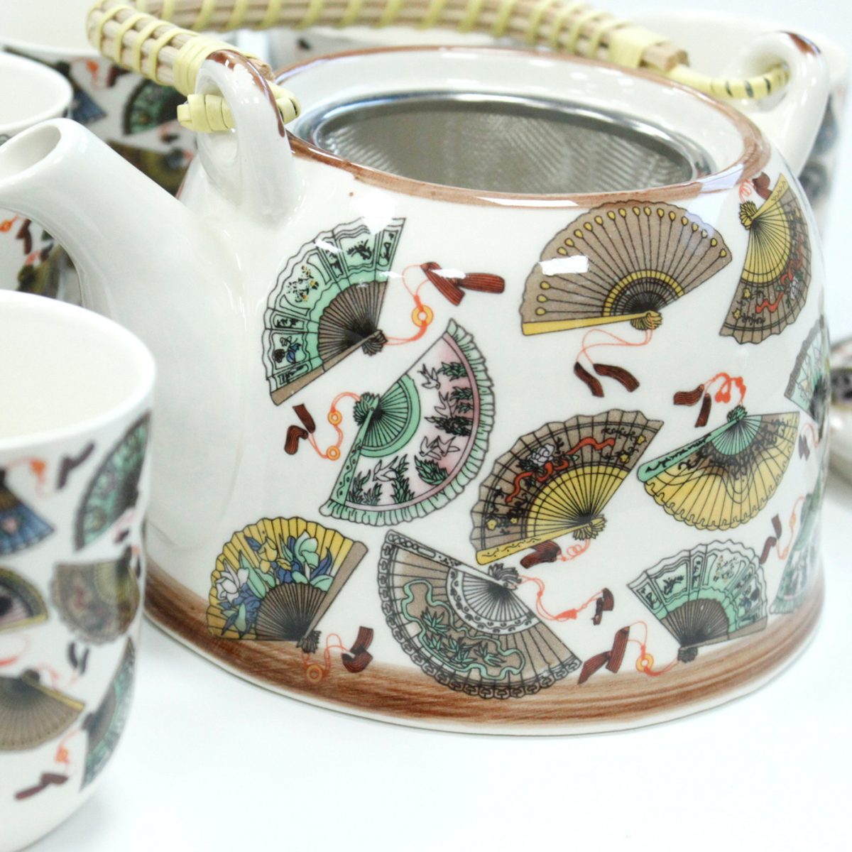 A ceramic teapot, adorned with colorful fan designs, sits on a white surface. It has a woven handle and is surrounded by similar patterned teacups, creating an ornate tea set.
