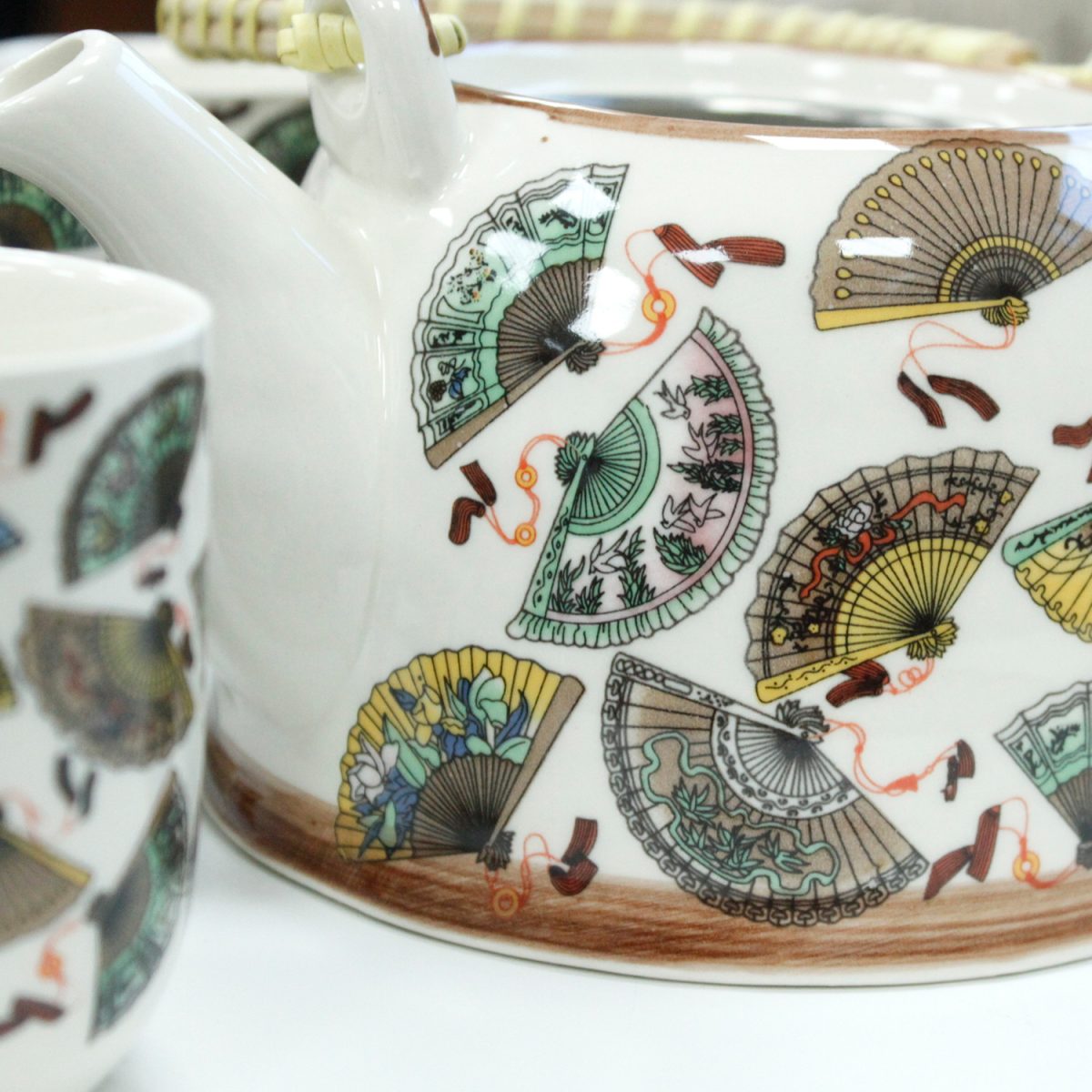 A white teapot and cup adorned with colorful, detailed fan designs. The teapot's bamboo handle and the intricate patterns create a decorative and elegant appearance. They rest on a light surface.