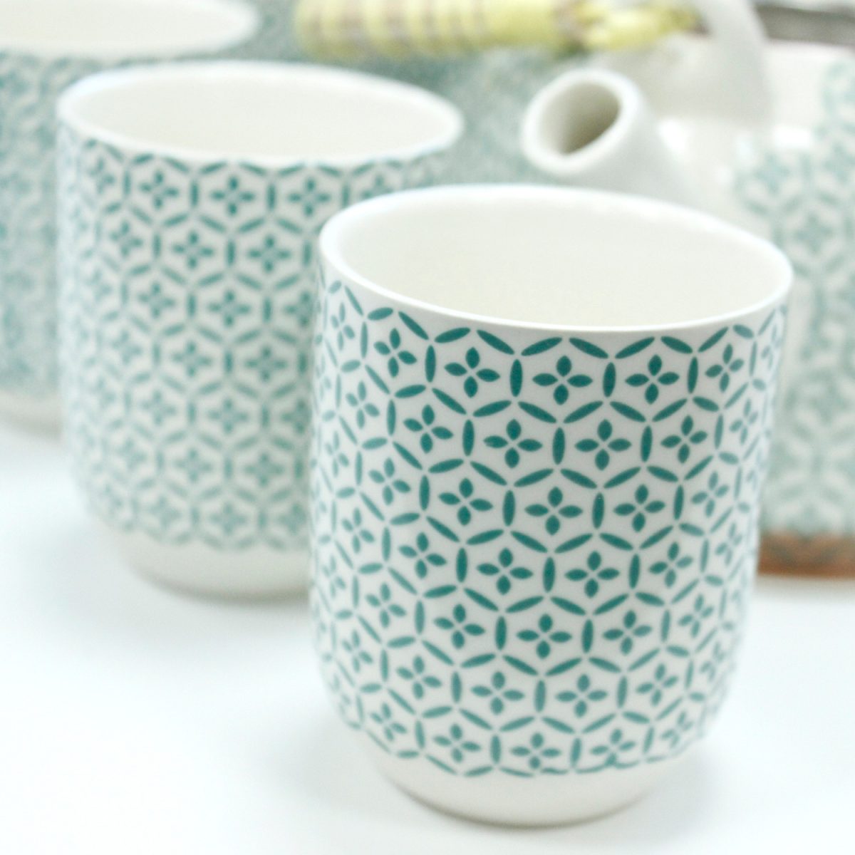 Ceramic cups feature a teal geometric pattern, standing stationary in a kitchen setting. A teapot is partially visible in the background, alongside other similar cups.