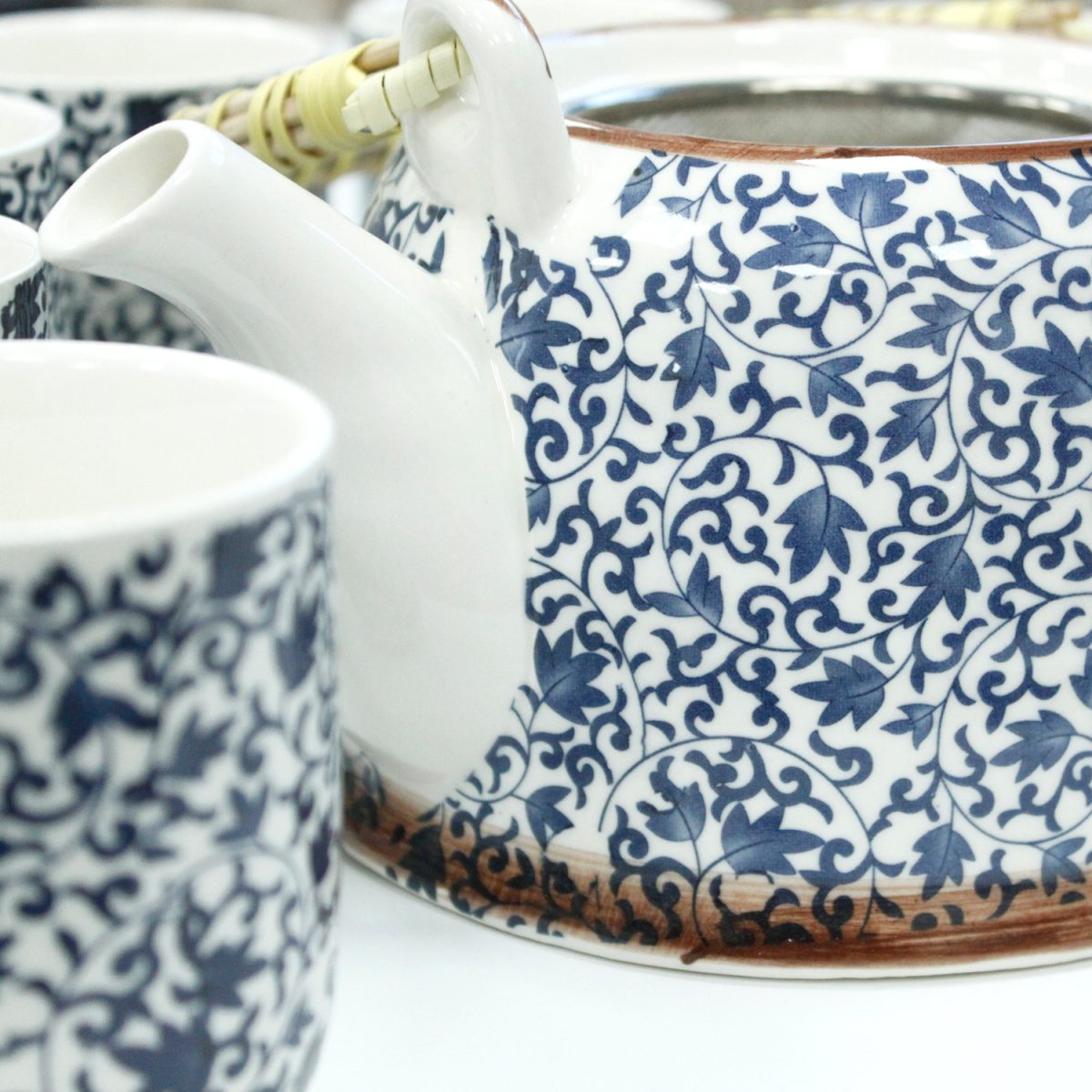 Teapot with white and blue floral pattern. Surrounded by matching cups, its spout faces left. Cane-wrapped handle suggests use in serving tea. Set on a light background.