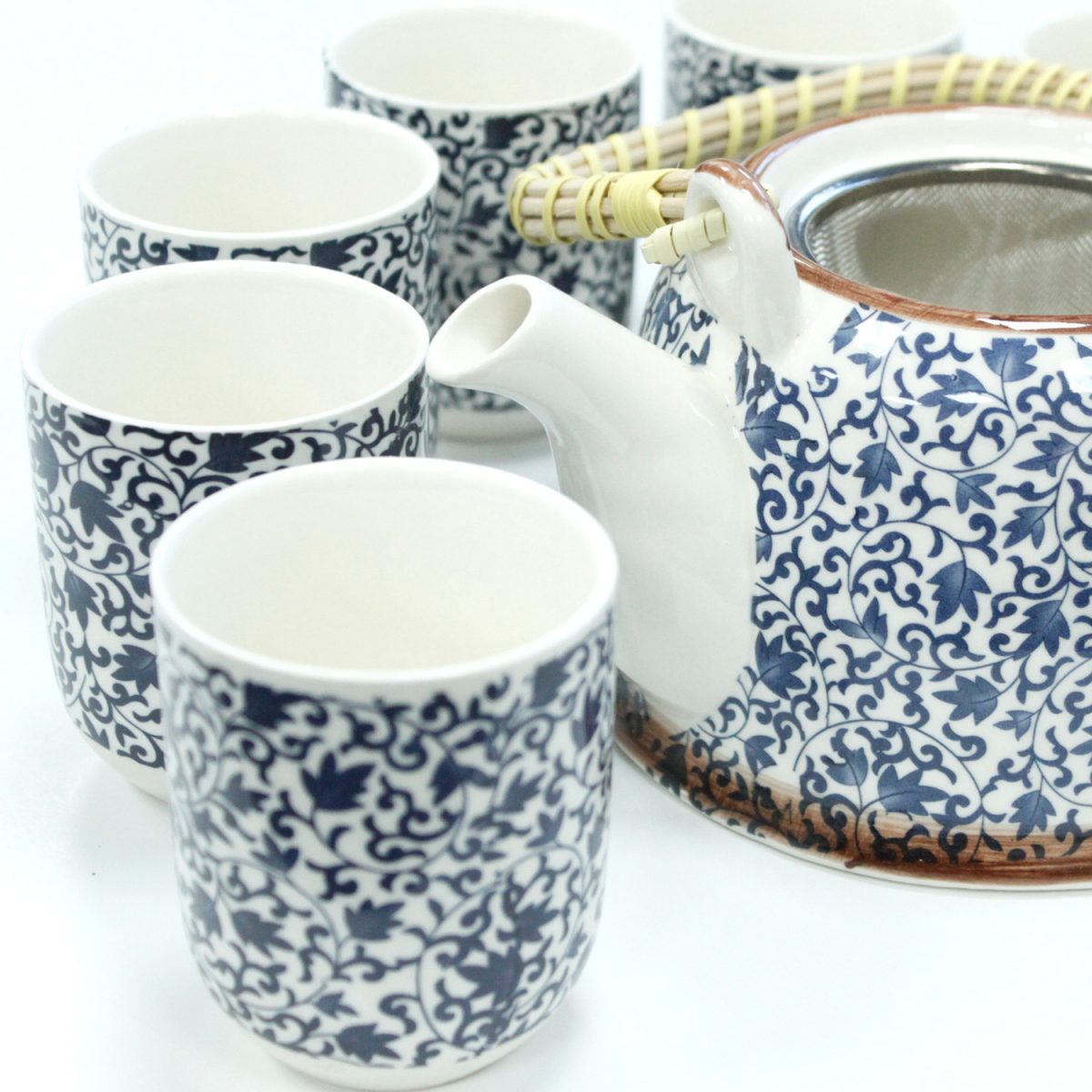A ceramic teapot, featuring blue floral patterns and a wicker handle, sits surrounded by six matching cups, creating an elegant tea set arrangement on a white background.