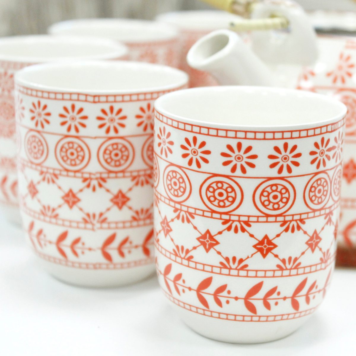 Ceramic cups, adorned with intricate red floral and geometric patterns, are arranged on a white table. A white teapot with a yellow handle is partially visible in the background.