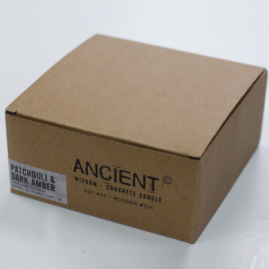 Cardboard box labeled "ANCIENT ©" sits on a white surface; front reads "WISDOM - CONCRETE CANDLE" and "SOY WAX + WOODEN WICK" and a sticker says "PATCHOULI & DARK AMBER".