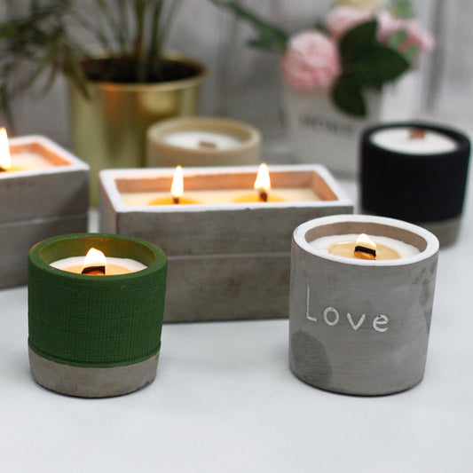 Concrete candle holders glow: a gray cylindrical holder engraved "Love", a green one and rectangular holders burning small flames on a white tabletop with blurred plants and pink flowers behind.