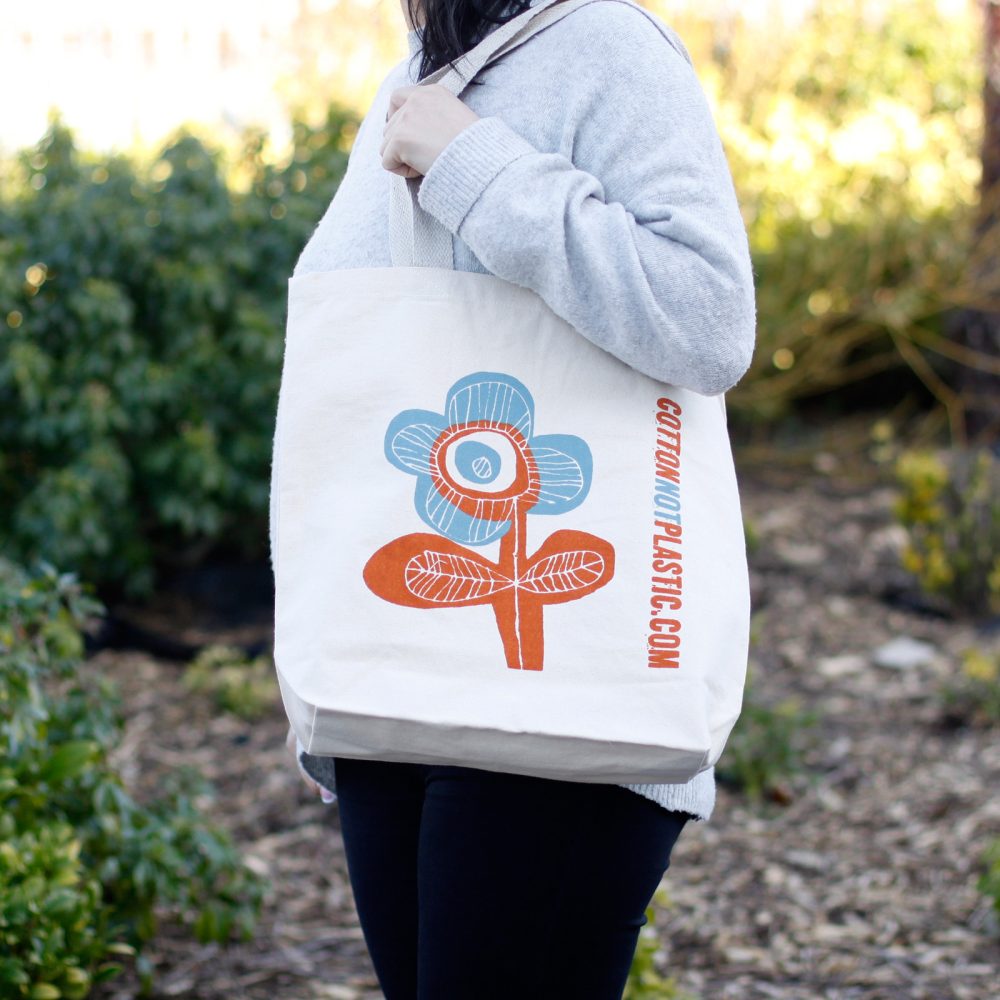 A person holds a tote bag with a large flower design and the text "COTTONNOTPLASTIC.COM." Bushes and trees stand in the background.