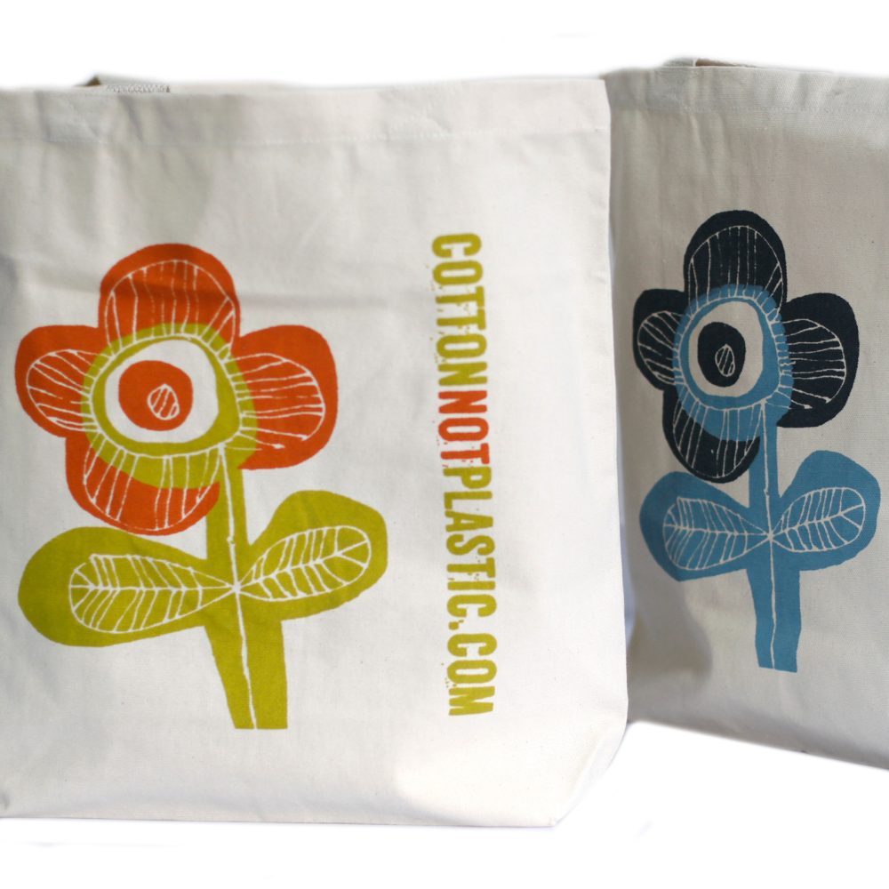 Two cream-colored tote bags display graphic flowers, one orange and green, and the other blue and black. Text reads "COTTONNOTPLASTIC.COM" vertically along the bag with the orange flower.