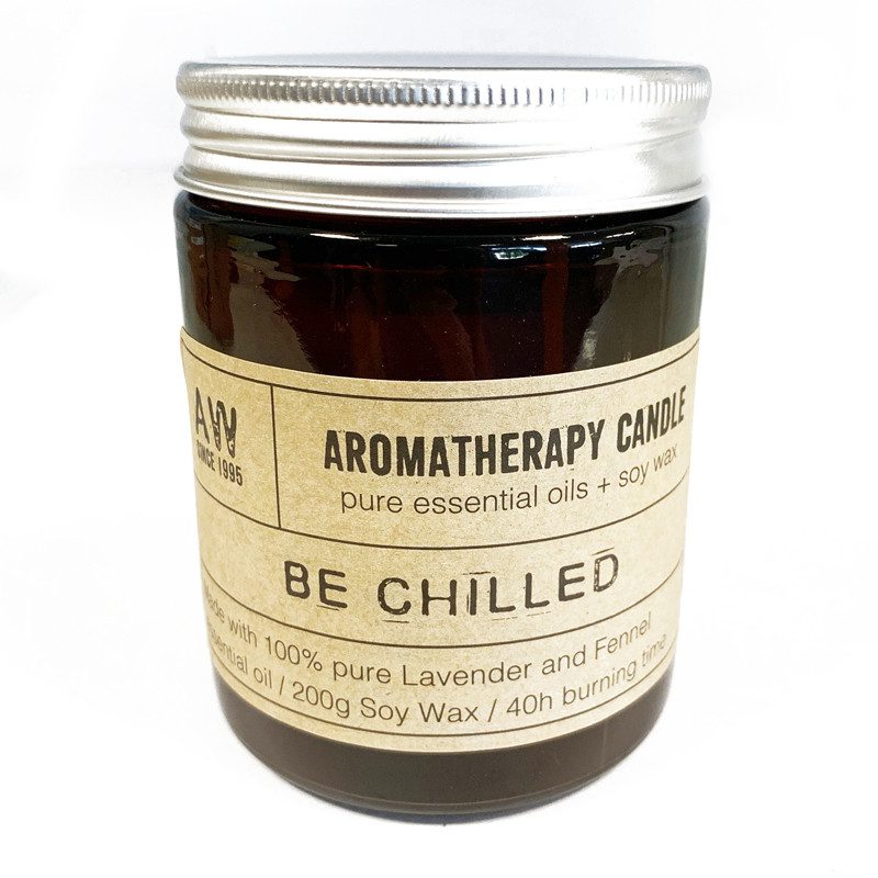 Aromatherapy candle in a dark glass jar with a silver lid sits on a white surface, featuring a label with text "AW SINCE 1995 AROMATHERAPY CANDLE pure essential oils + soy wax BE CHILLED de with 100% pure Lavender and Fennel sential oil / 200g Soy Wax / 40h burning time".