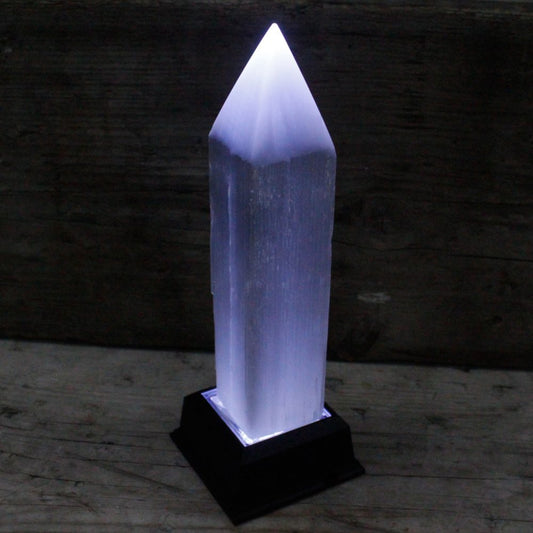 A selenite tower glows with internal light while sitting atop a black base. All of this is against a rustic wooden background.