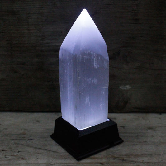 A glowing selenite tower sits atop a black, square base, illuminating upward. The background is a rustic wooden surface, both horizontal and vertical.