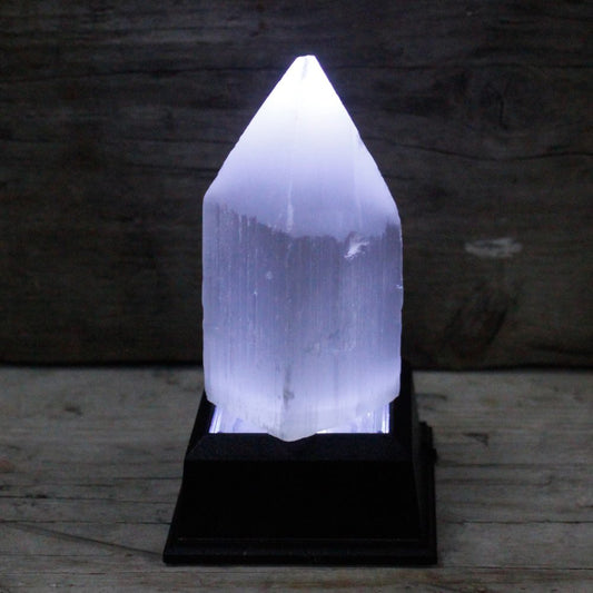 A glowing white selenite tower sits on a black base, emitting light. The background is a blurry wooden surface.