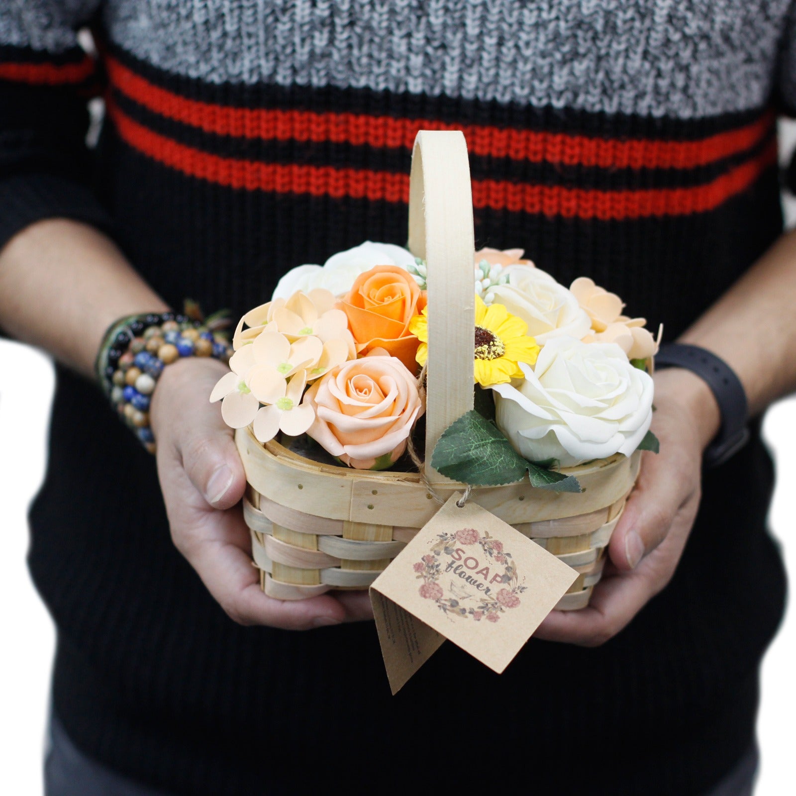 A person holding a small woven basket filled with various soap flowers, including roses and a sunflower. A tag is attached to the basket that reads "SOAP flower." The person is wearing a striped sweater.