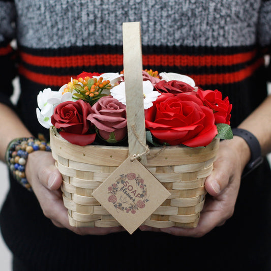 A woven basket holds a bouquet of red, pink, and white soap flowers labeled "SOAP flower," cradled by hands wearing bracelets and a striped sweater in a neutral background.