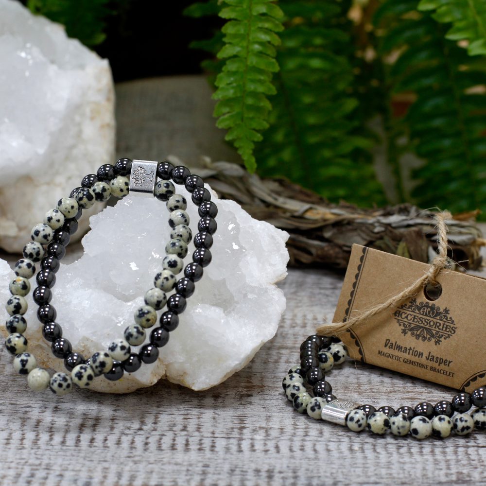 Two beaded bracelets rest on a quartz crystal, and another bracelet is next to a tag that reads "Accessories; Dalmatian Jasper Magnetic Gemstone Bracelet," on a wooden surface amid foliage.