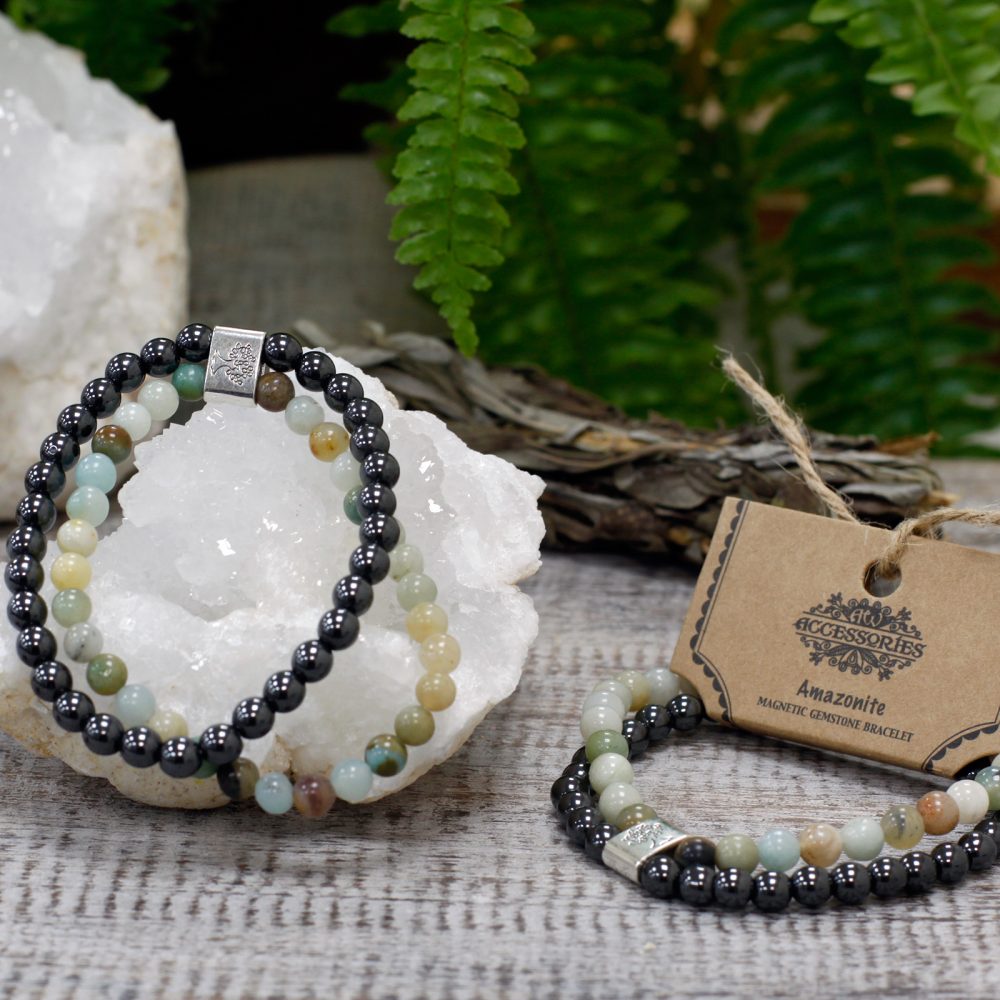 Two beaded bracelets, featuring black and earth-toned stones, rest on a white crystal near a tag that reads "Accessories, Amazonite, Magnetic Gemstone Bracelet," amidst foliage and wooden planks.