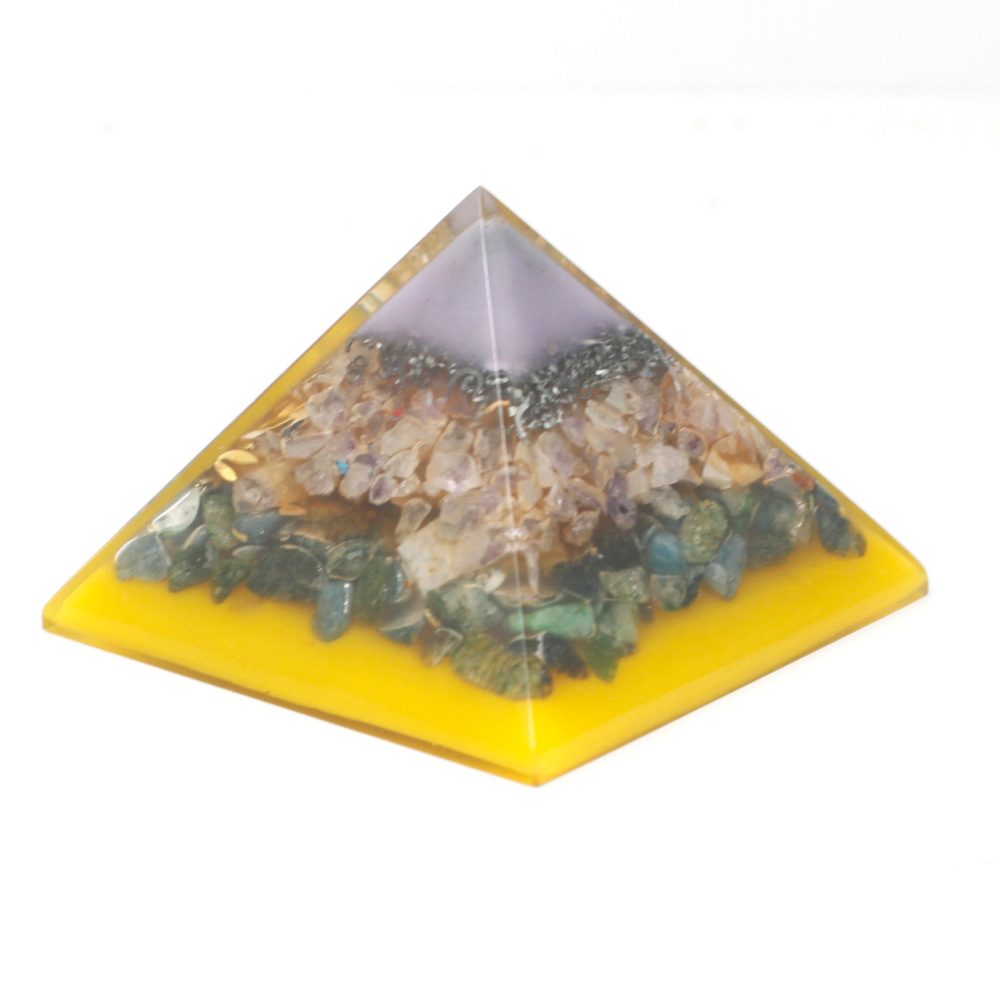 A layered orgone pyramid sits on a white surface. The base is yellow, filled with green and blue stones, then amethyst crystals, a layer of black stones, and a clear purple tip.