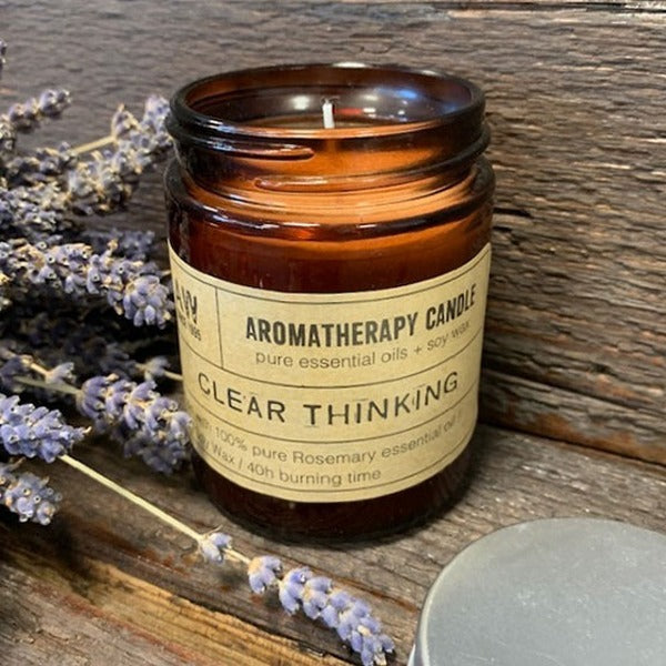 A brown glass jar candle, burning, sits on a rustic wooden surface near dried lavender.  The label reads: "AROMATHERAPY CANDLE pure essential oils + soy wax CLEAR THINKING 100% pure Rosemary essential oil Wax / 40h burning time".