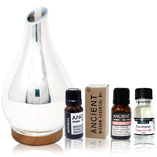 A silver glass diffuser sits on a wooden base, next to three small bottles of essential oils labeled "Happiness," "Grapefruit," and "Eucalyptus."