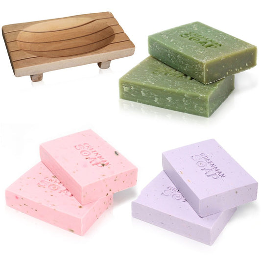 A wooden soap dish sits beside stacks of rectangular Greenman Soap bars in pink, lavender, and green.