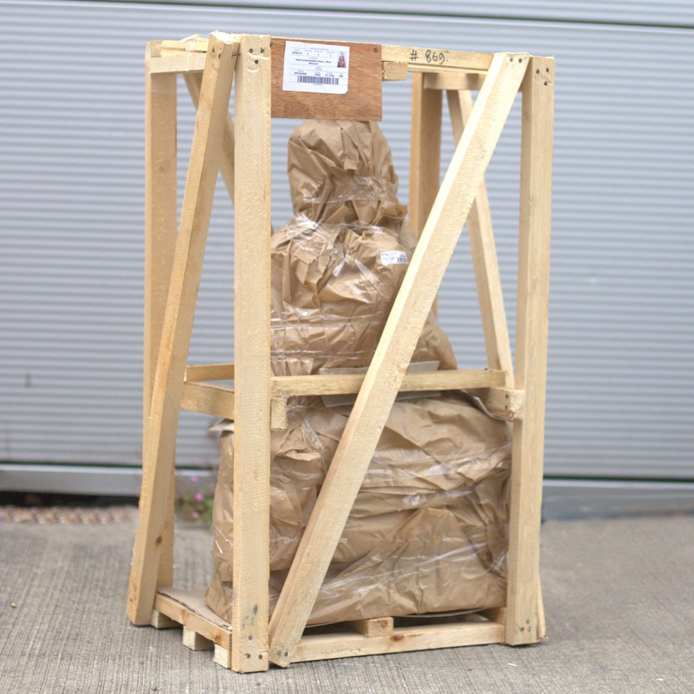 A wooden crate holds a large, brown paper-wrapped object, secured with tape. The crate sits on a concrete floor with a corrugated metal backdrop. A label reads, "#869".