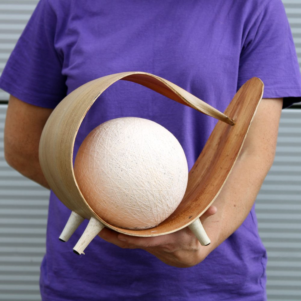 A person holds a wooden, curved stand cradling a large, off-white sphere.  The stand has small, light-colored legs. The context is outdoors against a corrugated metal wall.
