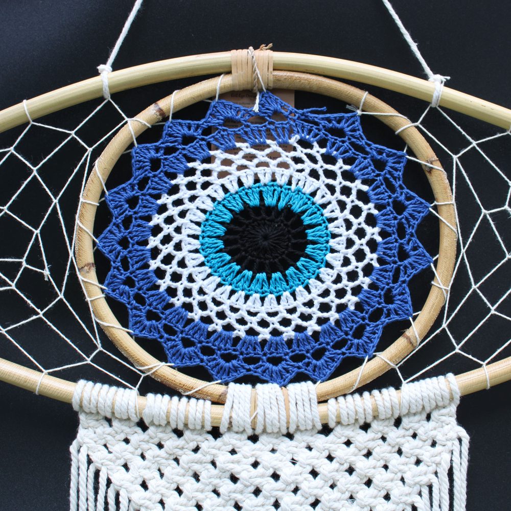 A crocheted evil eye hangs from a bamboo hoop, creating a dreamcatcher.  The woven design features blue, white, and turquoise against a black background.  It’s displayed against a dark backdrop.