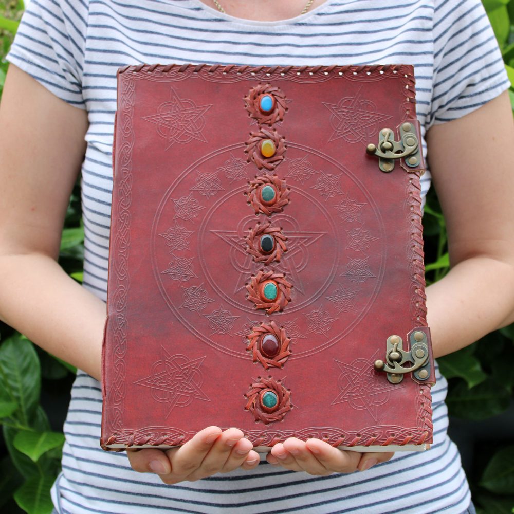 A person holds a large, red, leather-bound book with two clasps and a column of six colorful stones in an outdoor setting with green foliage. The book has patterns of stars and circles etched in.
