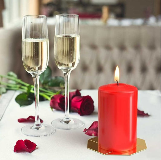 A red pillar candle burns softly on a white tablecloth beside two filled champagne flutes and scattered red rose petals in a cozy tufted chair dining setting.