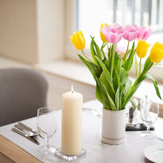 Bouquet of pink and yellow tulips sits in a white ceramic vase, accompanied by a lit ivory pillar candle and glassware on a set dining table by a sunlit window.
