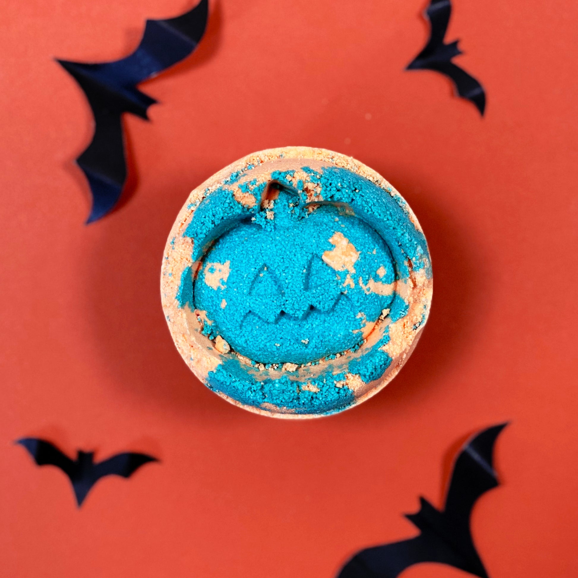 A blue pumpkin-shaped bath bomb rests on an orange surface, surrounded by decorative black paper bats, creating a Halloween-themed setting.