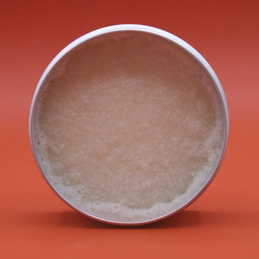 Round metal tin holds pale beige granular scrub, sits upright on a smooth bright orange surface against an orange background.