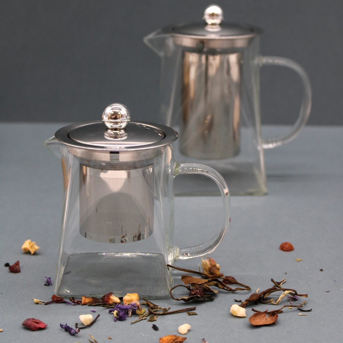 A clear glass teapot with a metal infuser and lid sits on a gray surface, surrounded by scattered dried tea leaves and petals. A similar teapot is slightly blurred in the background.