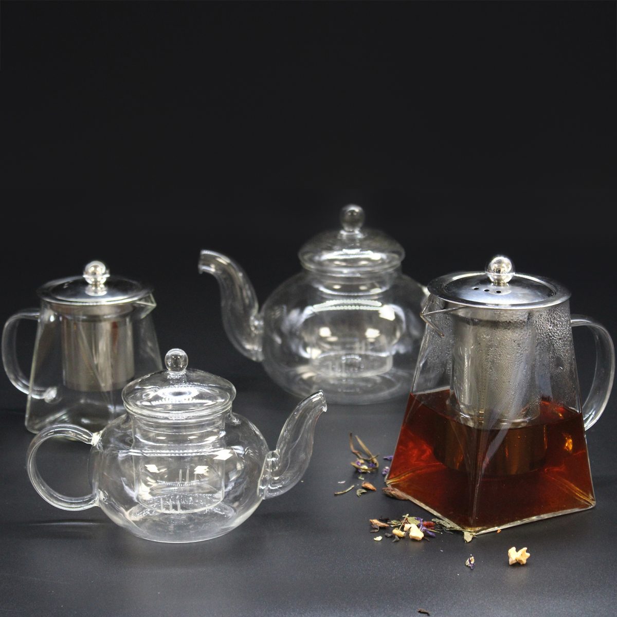 Glass teapots, some empty and one filled with tea, sit on a dark surface surrounded by scattered tea leaves under soft lighting.