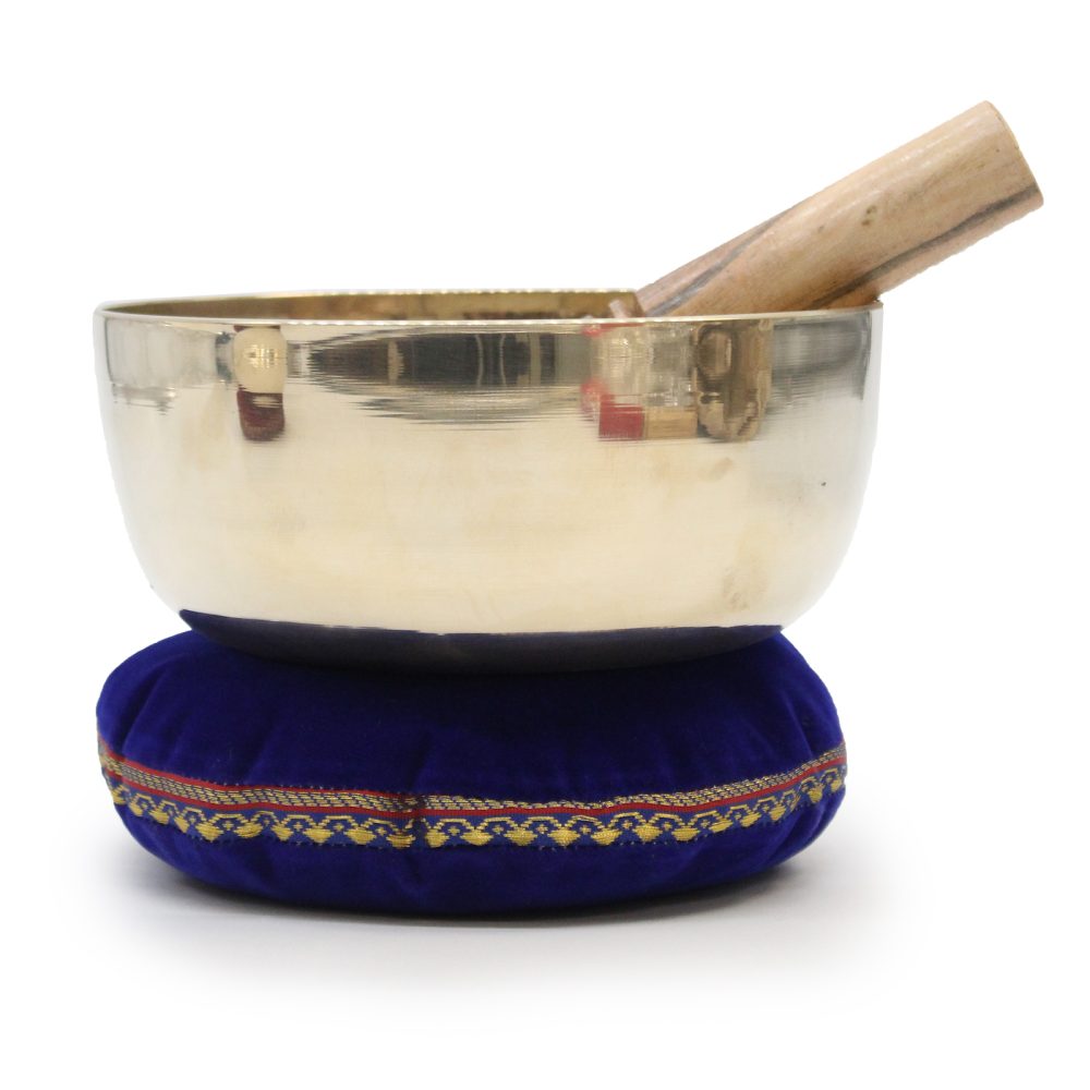 A metallic singing bowl rests on a decorative blue cushion with gold trim, accompanied by a wooden mallet inside, against a plain white background.