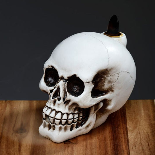 A white skull-shaped candle rests on a wooden surface. The wick is burned, emitting a slight stream of smoke. The background is a plain, dark gray.