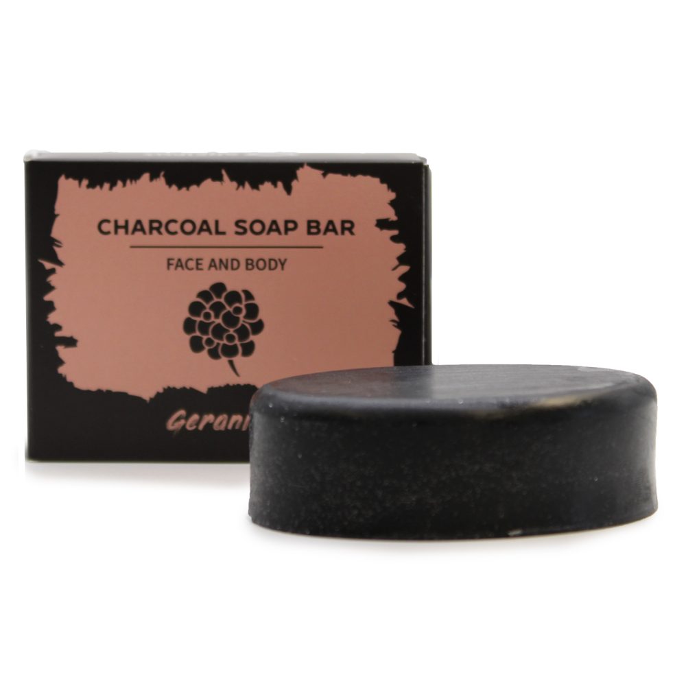 A round, black charcoal soap bar sits on a white surface, next to a black box labeled "CHARCOAL SOAP BAR FACE AND BODY", which also includes a floral design and the word “Gerani”.