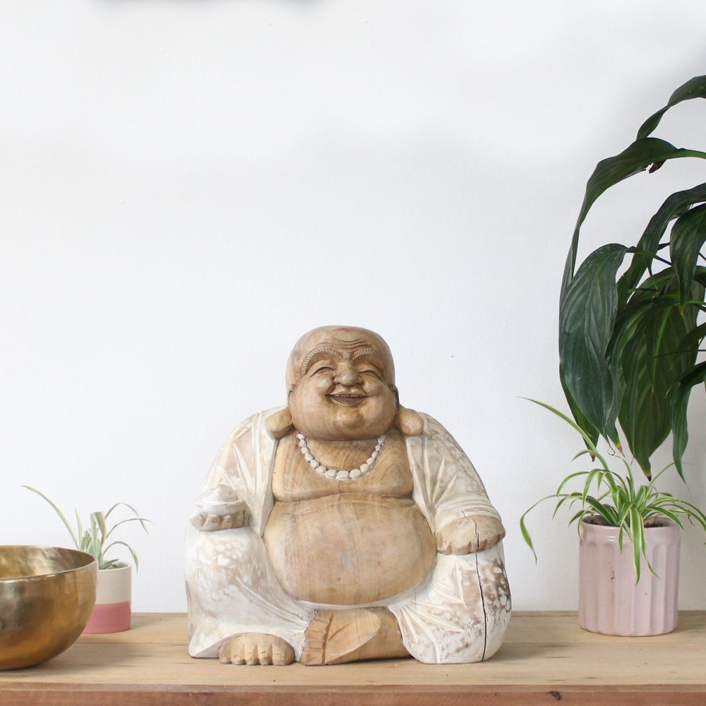 Wooden laughing Buddha statue sits serenely on a wooden surface. It is surrounded by a brass bowl and two small potted plants, all set against a plain white wall.