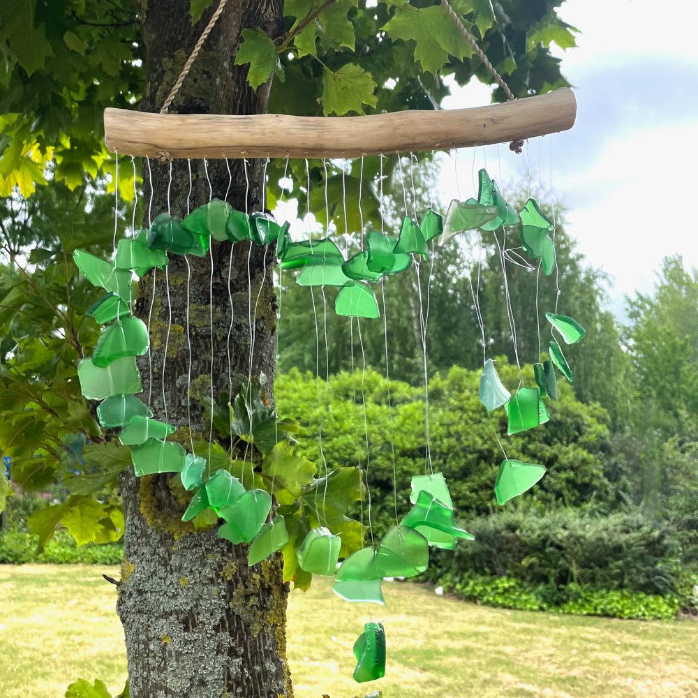 A wind chime, crafted from numerous green glass pieces, hangs from a driftwood branch.  It gently moves in an outdoor setting, near a tree, within a grassy lawn and lush green foliage.