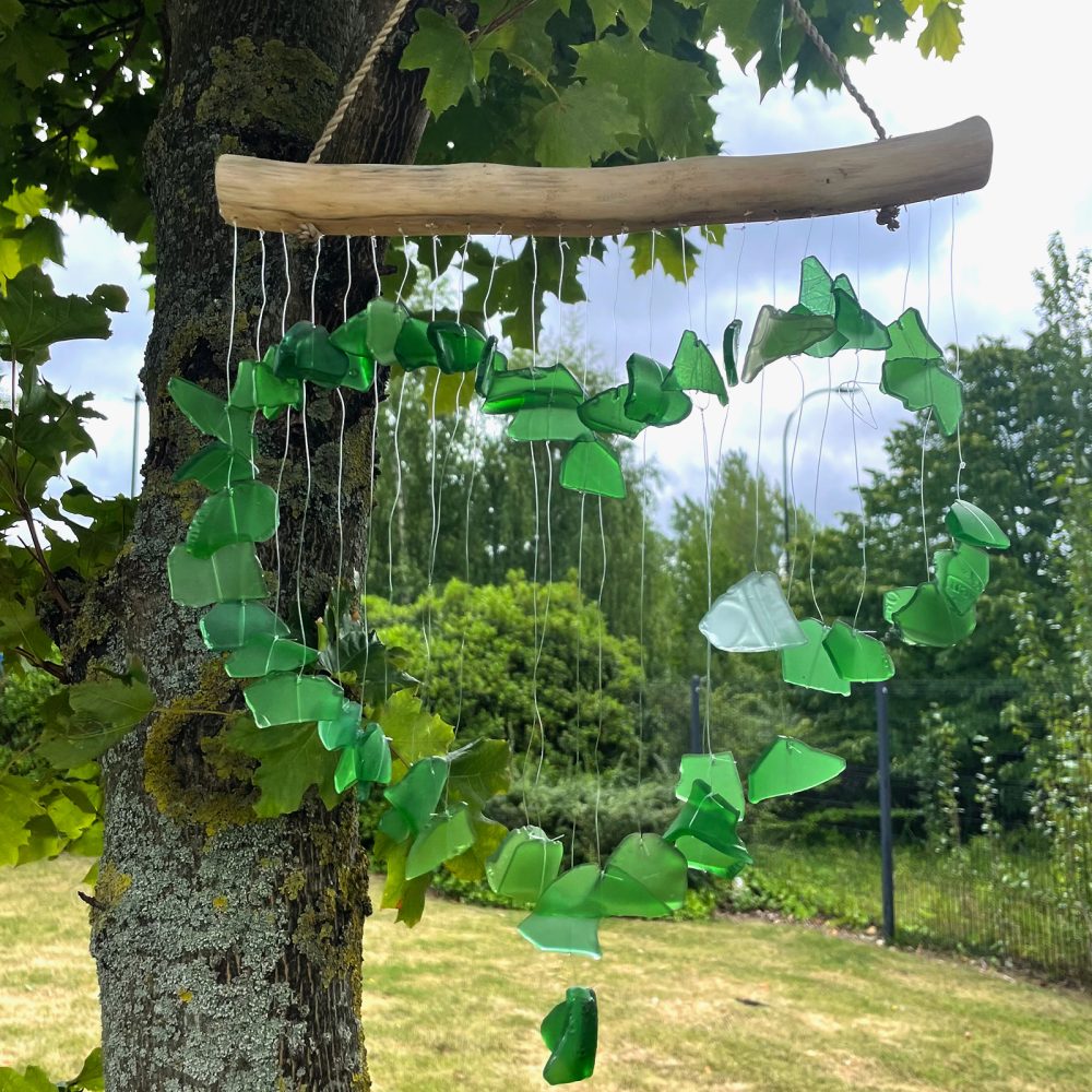 A heart-shaped wind chime, made of green glass pieces, hangs from a wooden branch.  It gently moves in a leafy, outdoor setting.