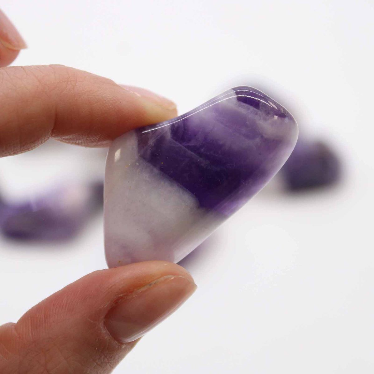A hand holds up a tumbled amethyst stone, revealing its polished surface, dark and light purple hues, and white patches. Other amethyst stones sit on the stark white backdrop.